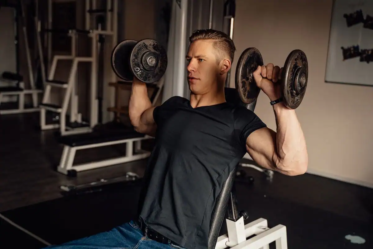 A man pressing a dumbbell overhead during a shoulder press in a gym
