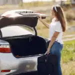 A woman holding a black rolling suitcase beside the open trunk of a silver car, about to load it for a road trip
