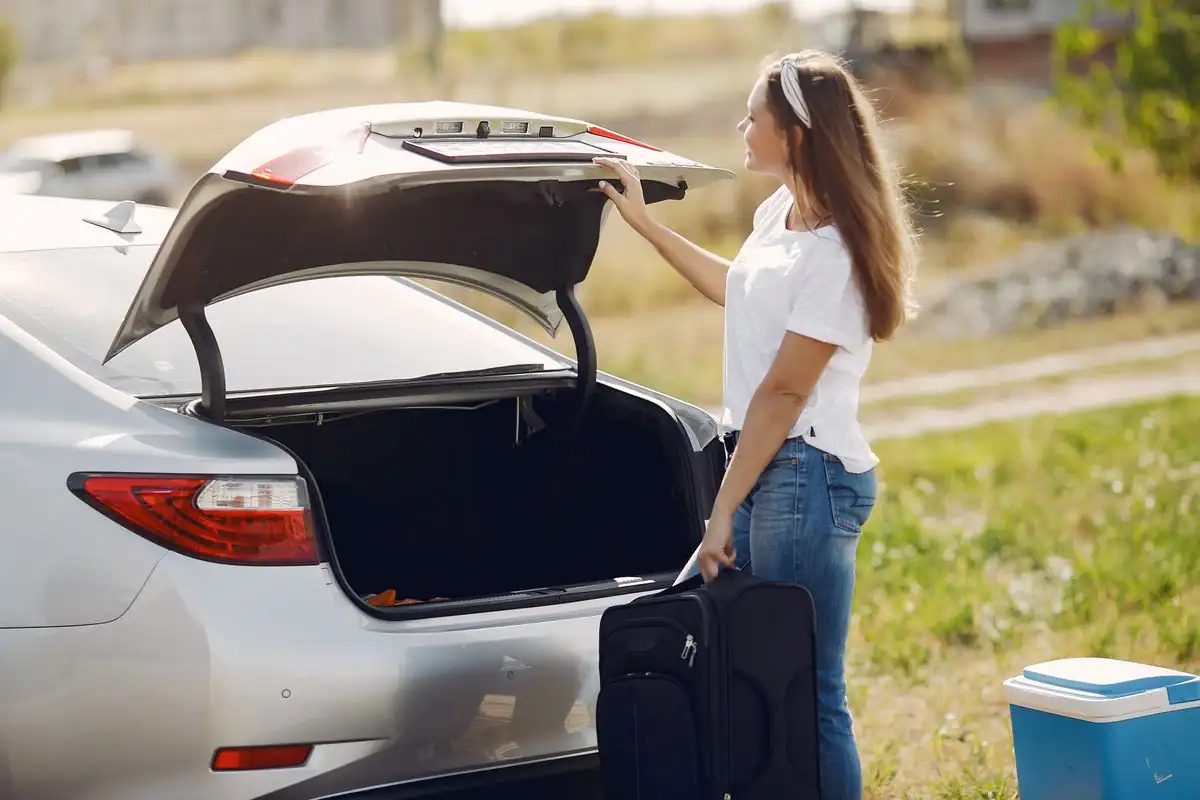 A woman holding a black rolling suitcase beside the open trunk of a silver car, about to load it for a road trip