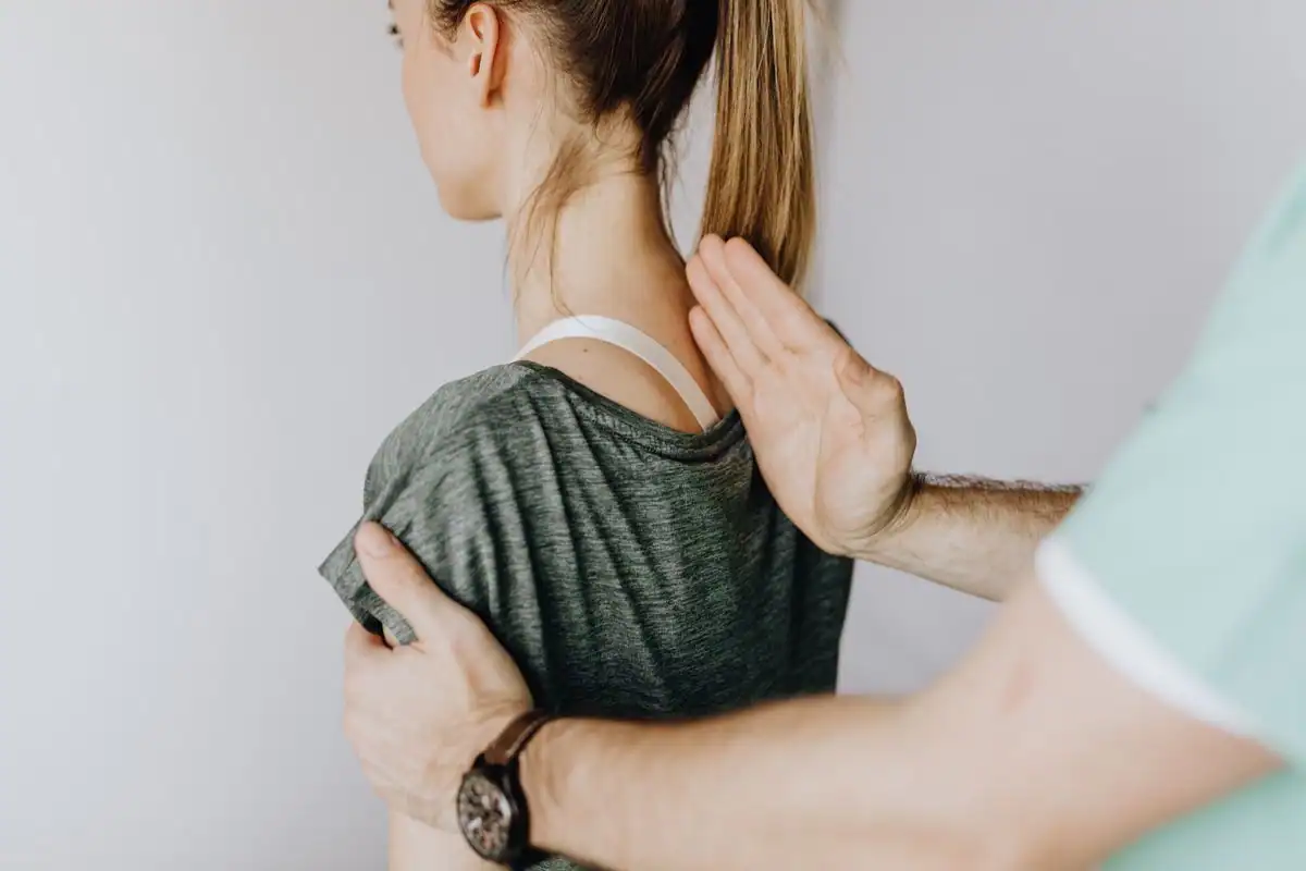 A chiropractor places one hand on a woman's upper back and the other on the front of her shoulder while checking her posture