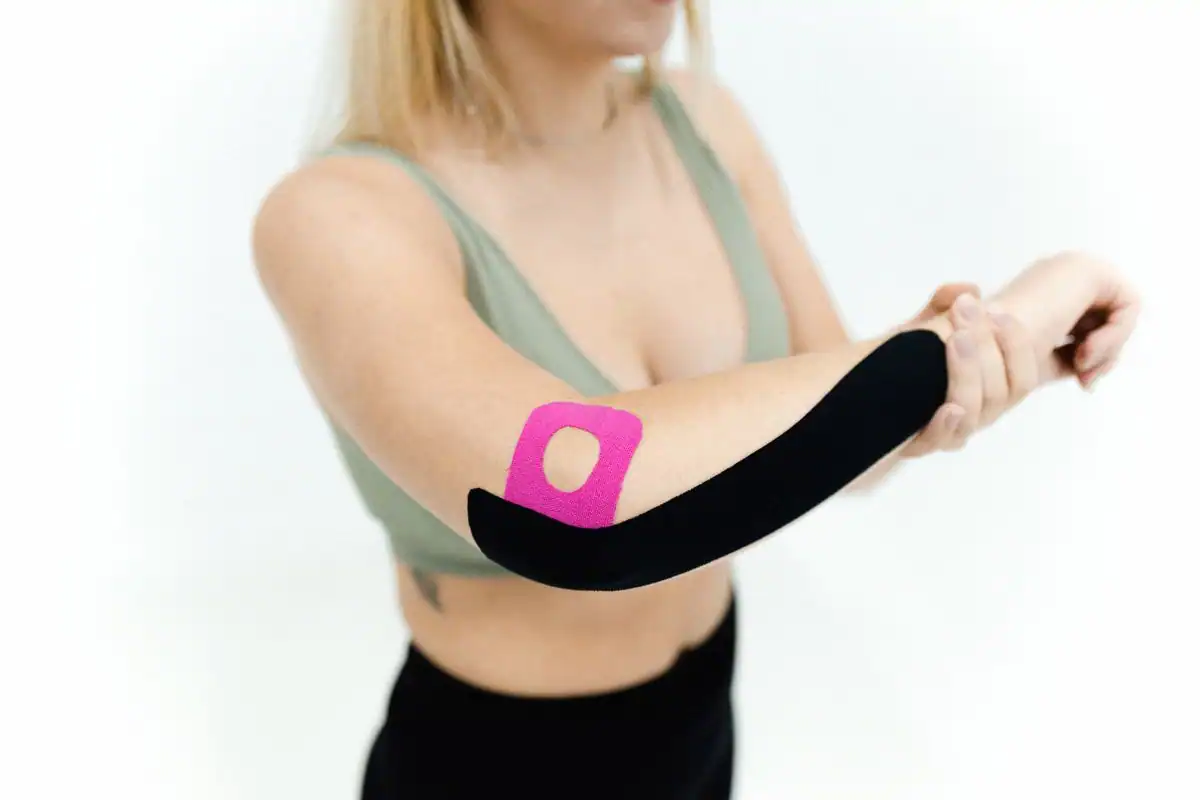 Woman stretching her wrist with pink and black kinesiology tape running along her outer elbow and forearm, a common setup for tennis elbow relief
